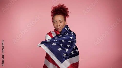 Smiling African American patriot redhead curly woman drapes US flag over shoulders. Portrait positive female standing with USA flag on isolated pink background. 4th July America independence day