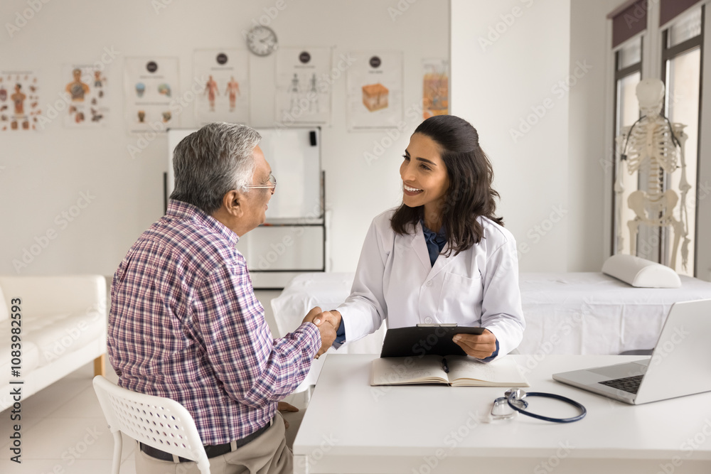 Obraz premium Friendly Latin female cardiologist in white coat shaking hands, greets old Indian male clinic patient, sit at desk, start medical consultation to listen health complains or disease symptoms. Eldercare