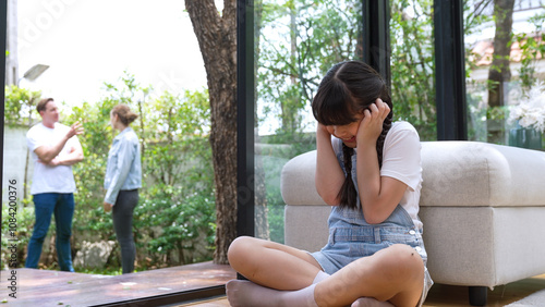 Stressed and unhappy young girl huddle in corner, cover her ears blocking sound of her parent arguing in background. Domestic violence at home and traumatic childhood develop to depression. Synchronos photo