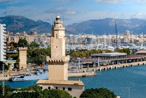 The Porto Pi lighthouse stands majestically with the Paraire’s tower in the background, creating a stunning panoramic view. In the foreground, a military ship sails through the calm waters.