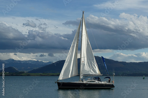 A sailboat is sailing through the bay of Santander, with the gentle waves of the Cantabrian Sea reflecting the daylight. The image captures the tranquility and elegance of sailing, as the boat moves a