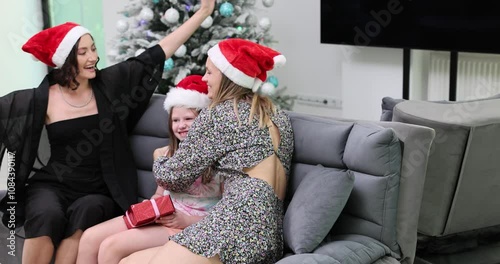 Young lesbian family in Santa hats sits on sofa during Christmas Eve. Daughter joyfully receives bright New Year gift from mother and aunt