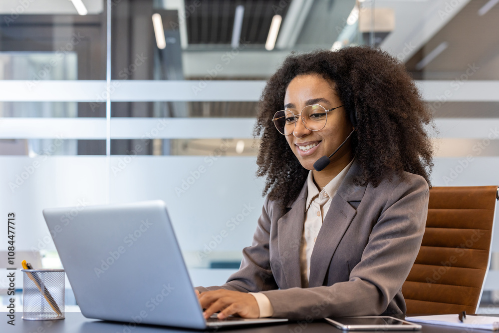 Smiling young African American business woman in a suit sitting at a desk and working on a laptop in the office Smiling young African American business woman in a suit sitting at a desk and working on a laptop in the office