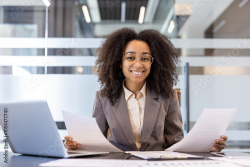 Portrait of a smiling young African American woman in a suit sitting at a desk in the office, holding documents and papers in her hands and looking at the camera