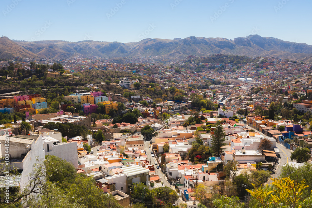 Fototapeta premium A panoramic view of vibrant Guanajuato, Mexico, with colorful houses and a sprawling cityscape surrounded by hills under a sunny sky.