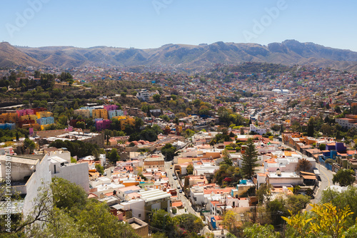 A panoramic view of vibrant Guanajuato, Mexico, with colorful houses and a sprawling cityscape surrounded by hills under a sunny sky.