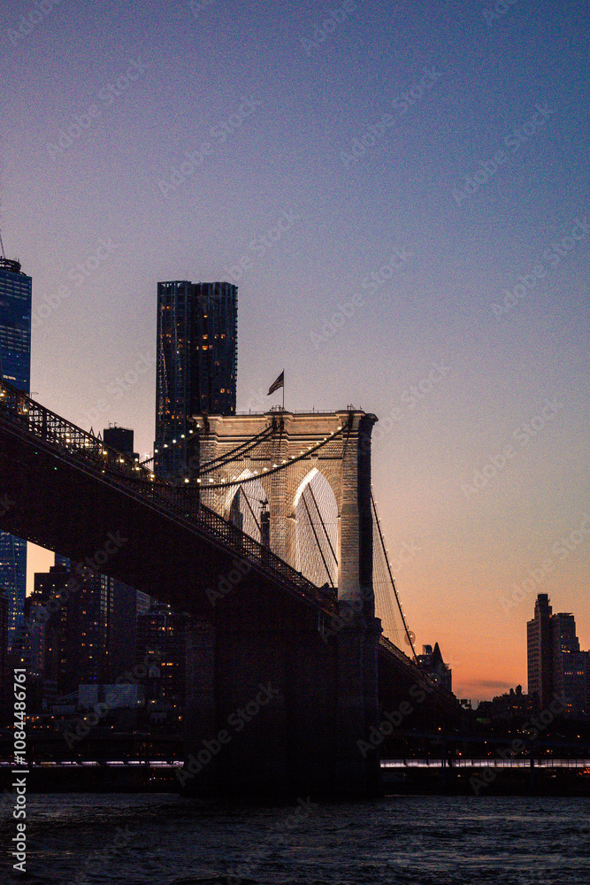 Obraz premium Elegant view of the Brooklyn Bridge illuminated at dusk, set against a soft gradient sky as the city skyline emerges in the background