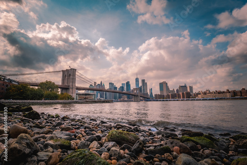 Scenic waterfront view of the Brooklyn Bridge and Manhattan skyline, with a rocky shoreline in the foreground and dramatic clouds filling the vibrant blue sky