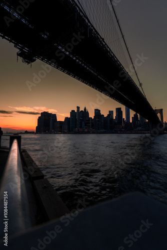 Silhouetted city skyline beneath the dramatic span of a bridge at dusk, with warm hues of the sunset reflecting on the tranquil waters.
