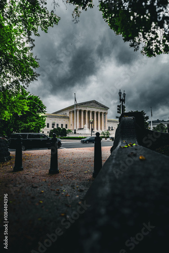 Moody outdoor scene featuring a neoclassical building with grand columns, framed by dramatic storm clouds and lush green trees, creating a striking contrast