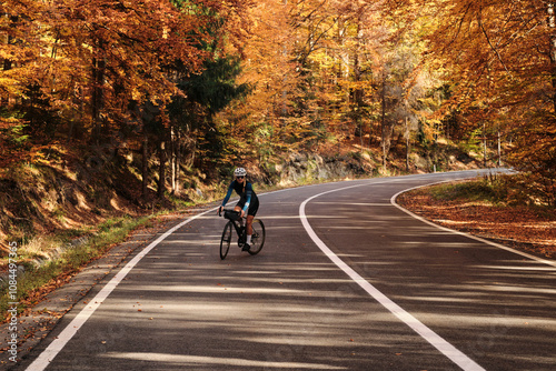 Woman cyclist rides on an asphalt road through a scenic autumn forest. Golden leaves create a vibrant backdrop for this active outdoor scene. Perfect for travel, sports, and nature themes.