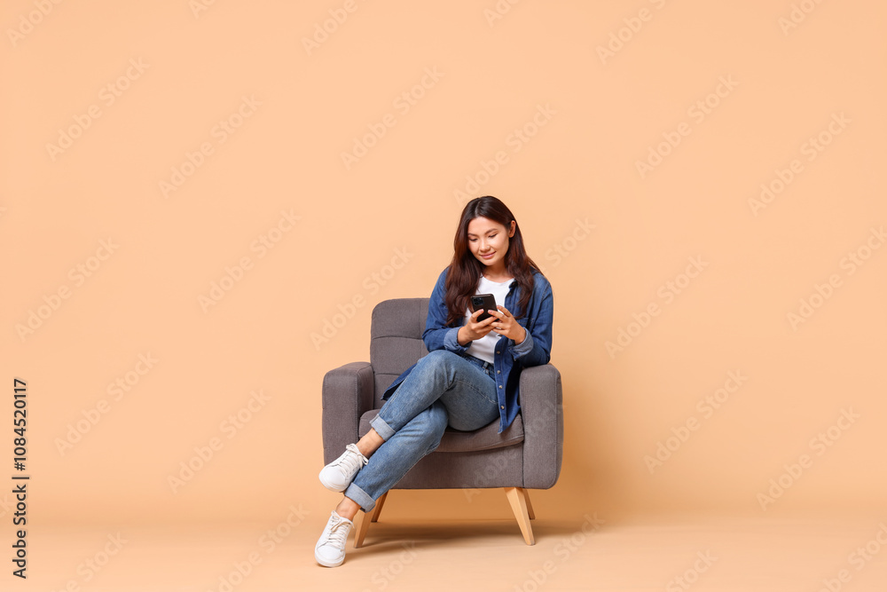 Young woman with smartphone sitting in armchair on beige background. Space for text