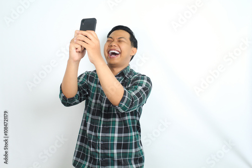 carefree asian man wearing casual plaid shirt laughing while watching video on smartphone isolated on white background