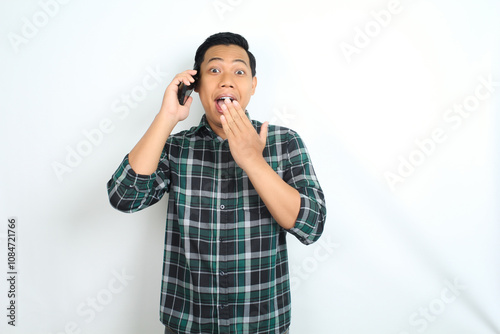 asian man covering mouth while talking on smartphone looks stunned isolated on white background