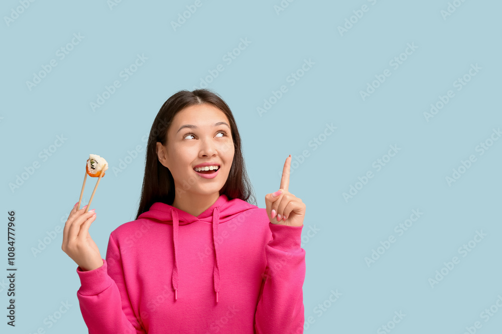 Beautiful young Asian woman holding chopsticks with tasty sushi roll and pointing at something on blue background Beautiful young Asian woman holding chopsticks with tasty sushi roll and pointing at something on blue background