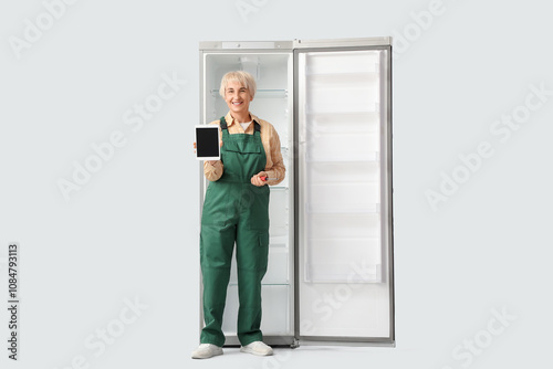 Female worker with tablet computer and refrigerator on light background