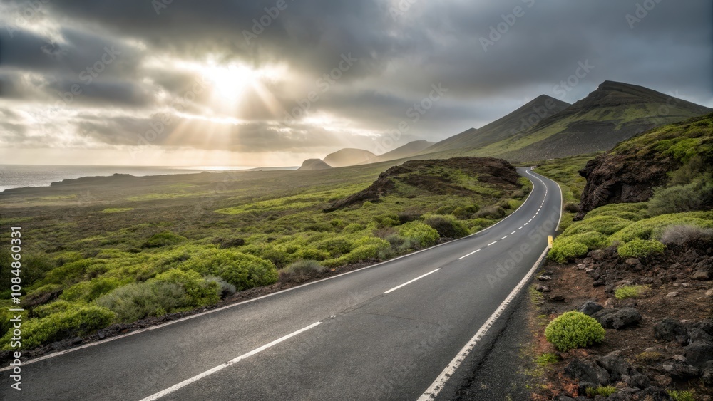 Fototapeta premium Asphalt road winding through a sun-drenched volcanic landscape with vibrant green shrubs and dramatic cloudy sky