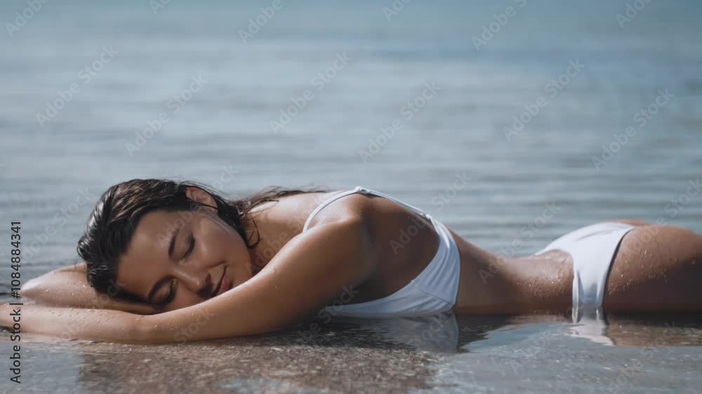 A woman in a white bikini lies peacefully on the beach, with waves gently washing over her. Perfect for themes of relaxation, summer, and serene beach moments. A woman in a white bikini lies peacefully on the beach, with waves gently washing over her. Perfect for themes of relaxation, summer, and serene beach moments.