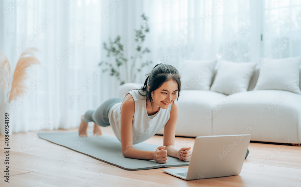 Young Asian woman in sportswear doing plank on training mat while watching online workout tutorials on laptop in the living room. Sport and recreation concept Young Asian woman in sportswear doing plank on training mat while watching online workout tutorials on laptop in the living room. Sport and recreation concept