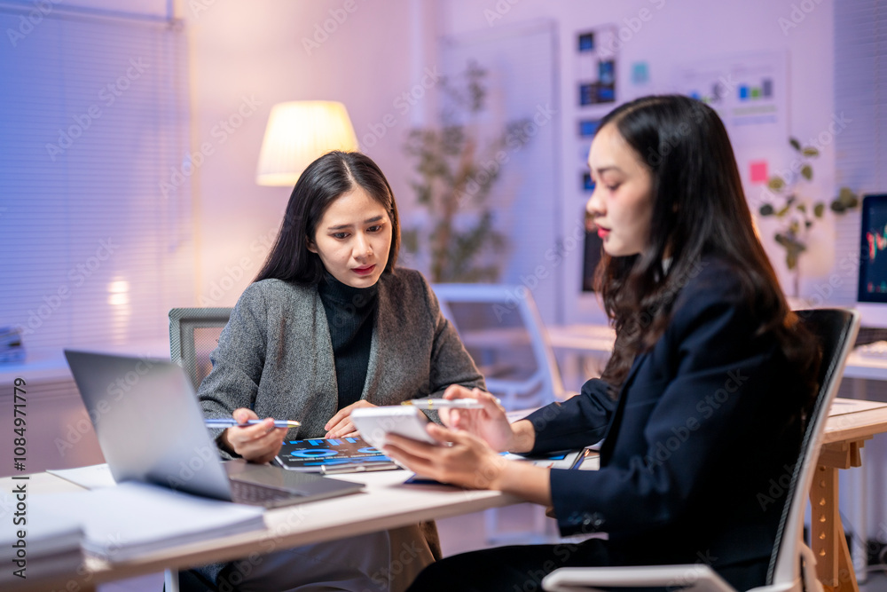 Two women are sitting at a desk, discussing something Two women are sitting at a desk, discussing something