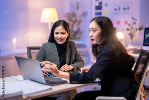 Two women are sitting at a desk with a laptop open in front of them