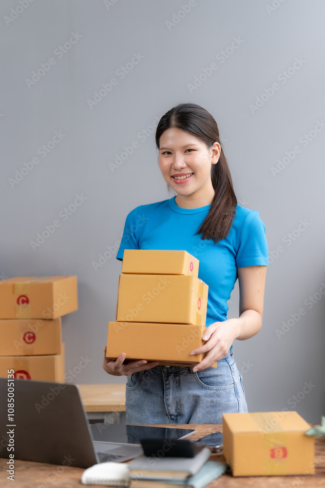 Smiling Woman Entrepreneur: A young woman in a blue t-shirt and jeans smiles confidently as she holds a stack of cardboard boxes in front of her. She stands in a home office setting. Smiling Woman Entrepreneur: A young woman in a blue t-shirt and jeans smiles confidently as she holds a stack of cardboard boxes in front of her. She stands in a home office setting.