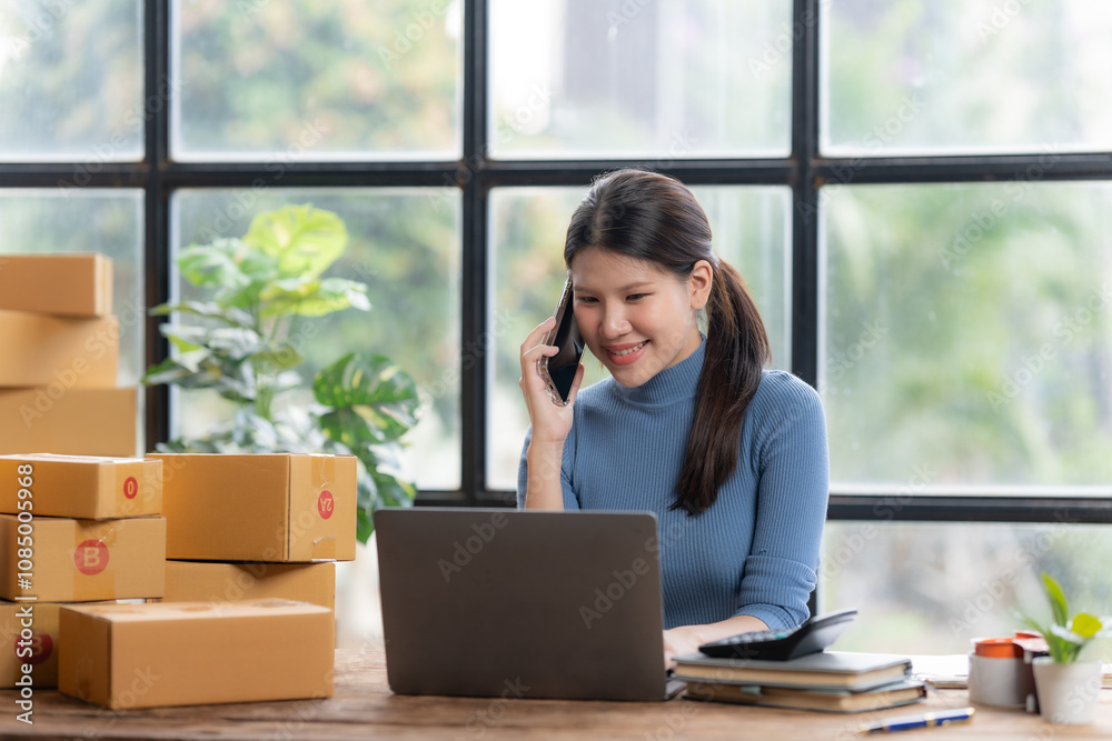E-commerce Success: A young woman manages her online business with a smile, taking a call while working on her laptop surrounded by boxes, showcasing the dynamism of modern entrepreneurship. E-commerce Success: A young woman manages her online business with a smile, taking a call while working on her laptop surrounded by boxes, showcasing the dynamism of modern entrepreneurship.