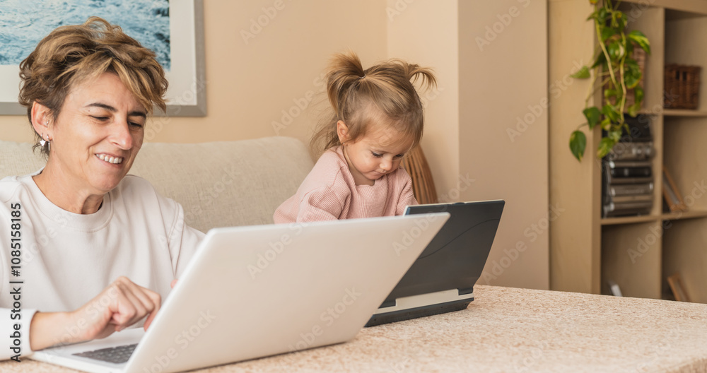 Woman And Little Girl Engaged In Work And Play With Laptops, Mature Woman Teleworking While Toddler Learns With Toy Computer, Family Bonding And Education Concept Woman And Little Girl Engaged In Work And Play With Laptops, Mature Woman Teleworking While Toddler Learns With Toy Computer, Family Bonding And Education Concept