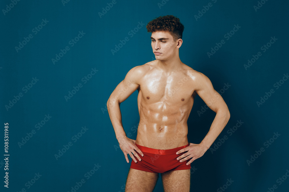 Confident young man posing in red swimwear, showcasing athletic physique against a vibrant blue background. Fitness and body image concept. Confident young man posing in red swimwear, showcasing athletic physique against a vibrant blue background. Fitness and body image concept.