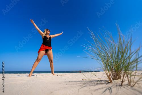 Young woman in one piece black swimsuit, orange pareo and sunglasses standing with her legs apart and her arms raised up on white sand on beautiful beach on sunny summer day. Energy. Front view