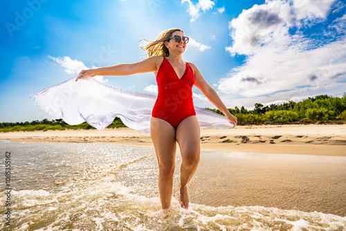 Summertime. Beautiful middle-aged woman in one piece red swimsuit holding white scarf fluttering in the wind, walking in shallow water on sandy beach by sea on, beautiful sunny day. Front view