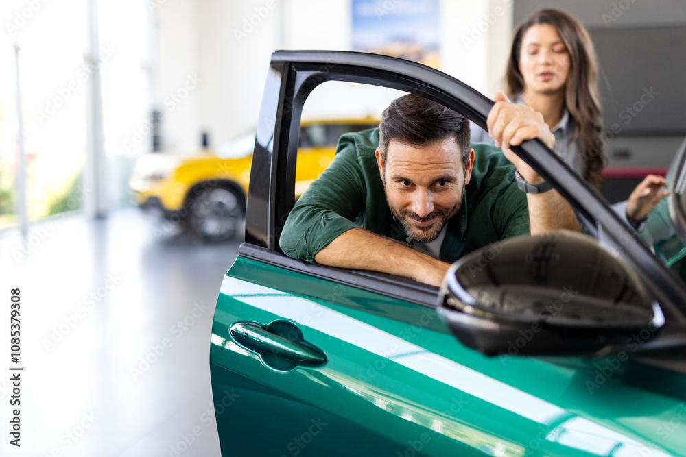 Happy young man hugging his new car in showroom. Satisfied guy with closed eyes embracing the hood of the automobile. Dreaming man lying on car bonnet hugging it. Happy young man hugging his new car in showroom. Satisfied guy with closed eyes embracing the hood of the automobile. Dreaming man lying on car bonnet hugging it.