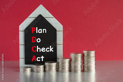PDCA symbol. Concept words PDCA Plan, Do, Check, Act written on a black board. Silver coins arranged in a graph in front. Beautiful red background, grey table. Business concept