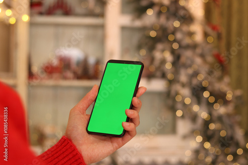 Green screen phone in a female hand. Gadget on the background of Christmas tree. Close-up of a female hand and a smartphone with a green screen, New Year. Modern mobile phone. Mockup. Technology