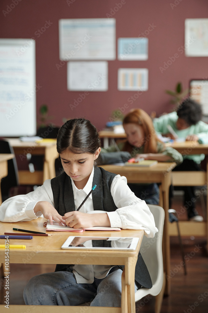 Diligent schoolgirl taking notes in copybook while sitting by desk against group of classmates at English grammar lesson
