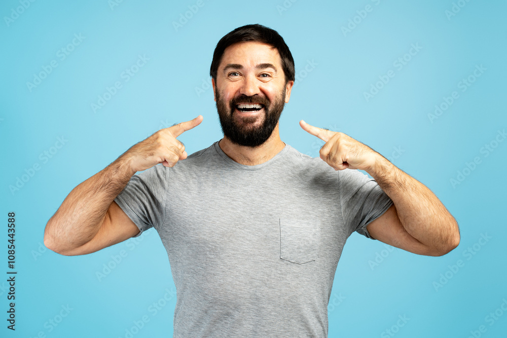 Smiling bearded man pointing his perfect white teeth with fingers on blue background Smiling bearded man pointing his perfect white teeth with fingers on blue background