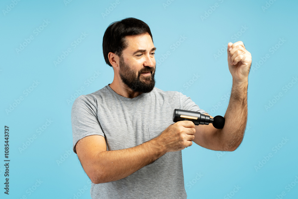 Man massaging his arm with a handheld massage gun on blue background Man massaging his arm with a handheld massage gun on blue background