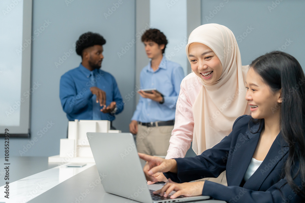 Two professionals working together on a laptop, exchanging ideas, while other team members discuss in the background of a modern office. Two professionals working together on a laptop, exchanging ideas, while other team members discuss in the background of a modern office.