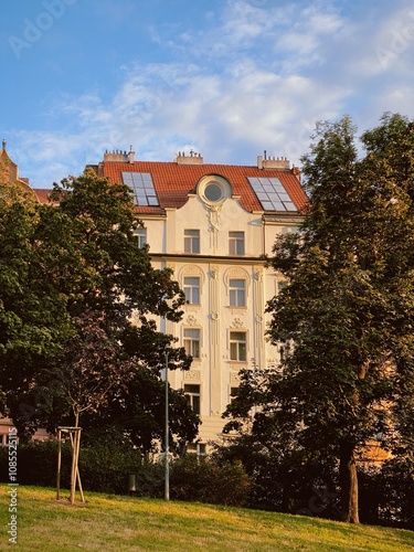 Golden hour view of a historic building in Prague's Vinohrady district, framed by lush green park trees and a vibrant blue sky. The intricate facade glows warmly in the evening sunlight.