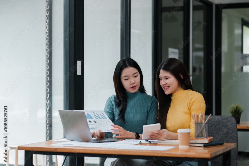 Two Young Women Collaborating in Modern Office Setting with Laptop and Documents for Business Strategy and Analysis Two Young Women Collaborating in Modern Office Setting with Laptop and Documents for Business Strategy and Analysis