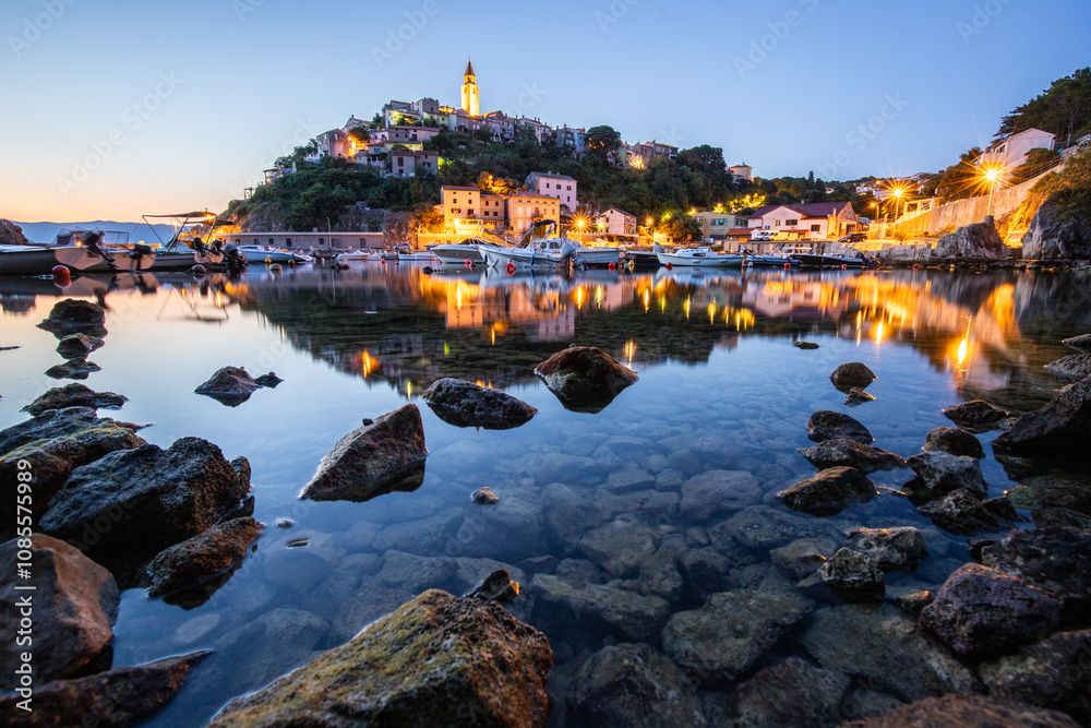 Harbor and beach with an old village on a hill. Boats are reflected in the water, the sun rises on the horizon of the Adriatic. Beautiful landscape shot from Vrbnik, Krk, Croatia Harbor and beach with an old village on a hill. Boats are reflected in the water, the sun rises on the horizon of the Adriatic. Beautiful landscape shot from Vrbnik, Krk, Croatia