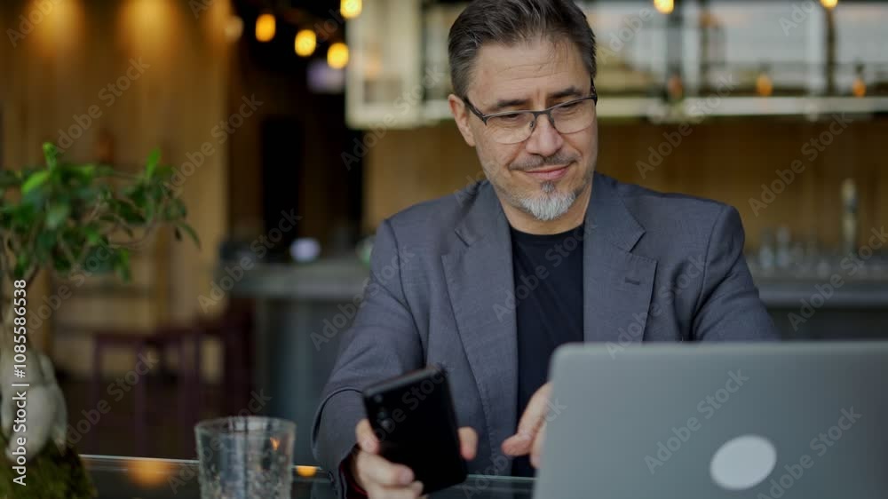 Businessman sitting at table in hotel lobby, restaurant or cafe, working on laptop computer. Mature age, middle age, mid adult man in business casual, confident happy smiling.