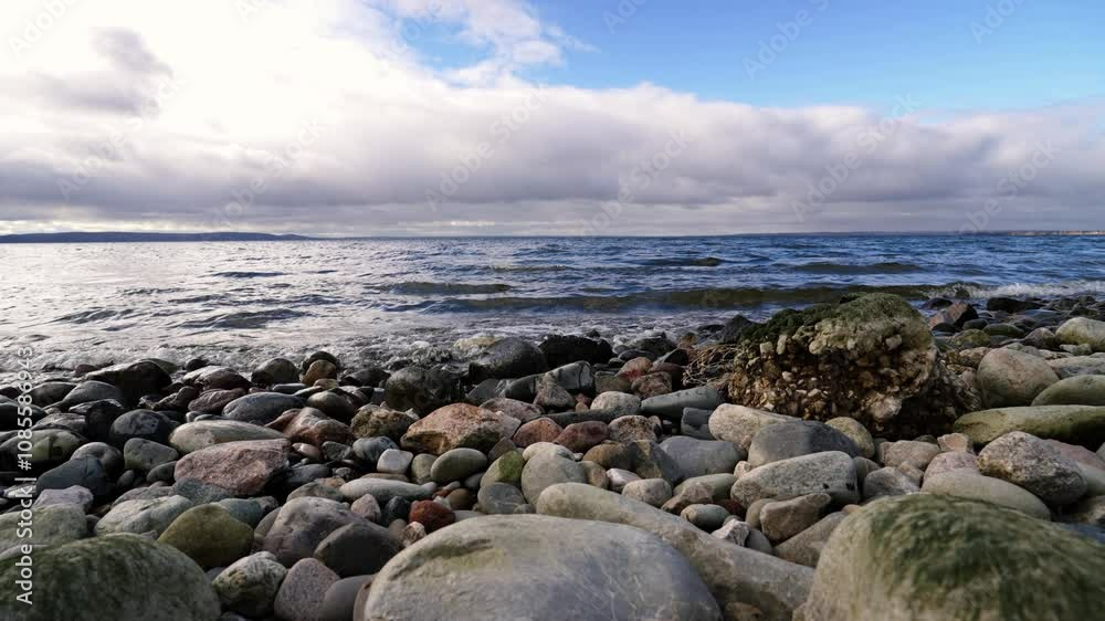 Ocean waves crash against a rocky beach under a bright sun, with white clouds drifting across a blue sky. The scene captures the beauty and tranquility of nature's coastline
