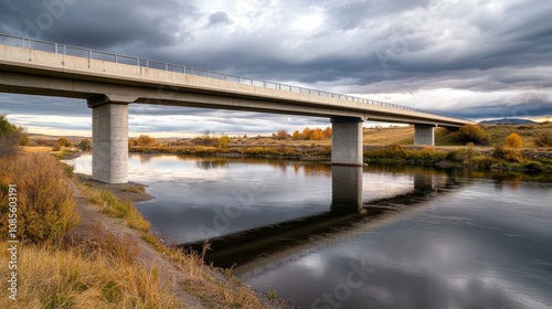 A scenic view of a bridge spanning a calm river, surrounded by lush vegetation under a cloudy sky, reflecting nature"s beauty.