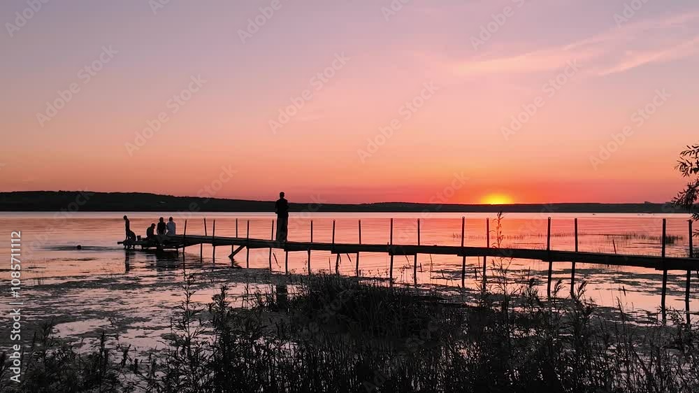 A colorful sunset over a lake with a pier, silhouettes of unrecognizable people walking along the pier. Slow motion video A colorful sunset over a lake with a pier, silhouettes of unrecognizable people walking along the pier. Slow motion video