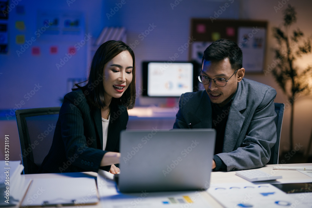 Fototapeta premium Two asian businesspeople are working together on a laptop in a modern office at night, collaborating on a project with documents and charts spread out on the desk