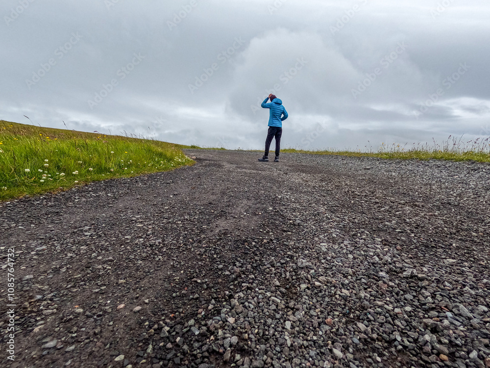 Rear view of a single female standing on a winding gravel road. She is dressed casually and holds a contemplative stance. Grassy field surrounds the road. Nólsoy, Faroe Islands.