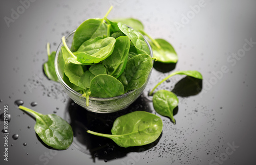 Spinach in a transparent glass bowl on gray background with reflection. Heap of Baby spinach fresh leaves close up, water drops, Healthy vegan food, diet, dieting, vitamin concept. 