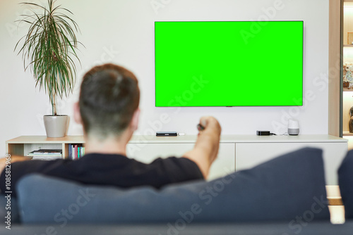 Rear view. The guy turns on the plasma flat-screen TV by pointing the remote at the green screen. The man is out of focus, watching TV while relaxing in his living room. Layout for the design.