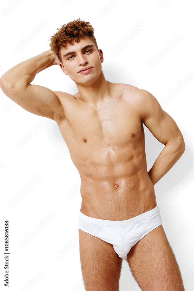Fototapeta premium Confident young man with curly hair posing against white background, showcasing athletic physique in minimal swimwear, representing health and fitness lifestyle.